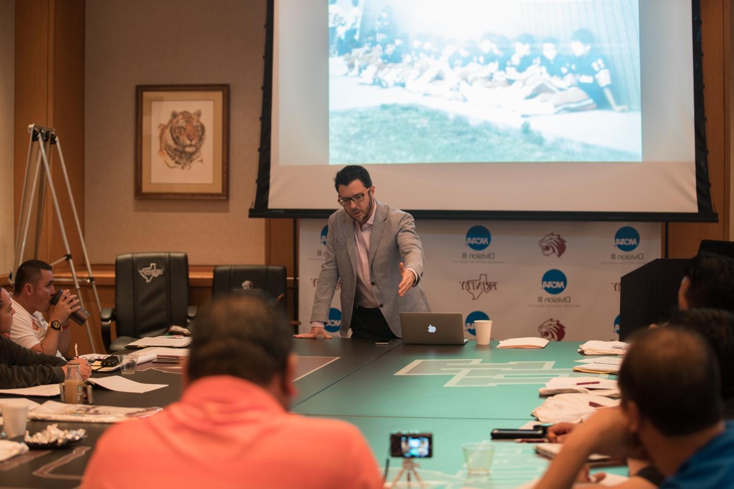 Speaker talking to attendees around a table with a projector screen behind him